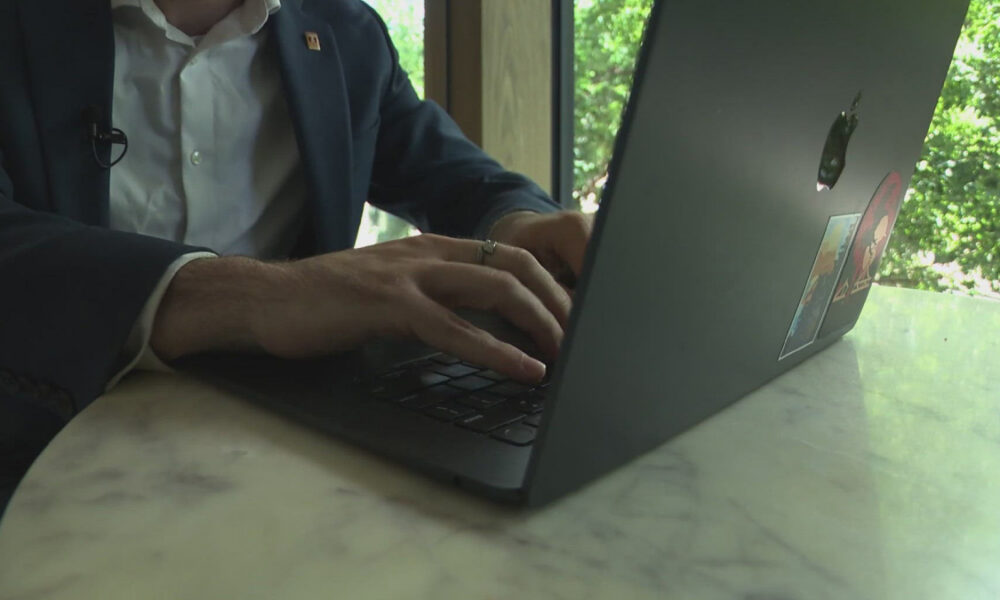 Person in a suit types on a MacBook with stickers at a marble table by a window with greenery outside