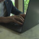 Person in a suit types on a MacBook with stickers at a marble table by a window with greenery outside