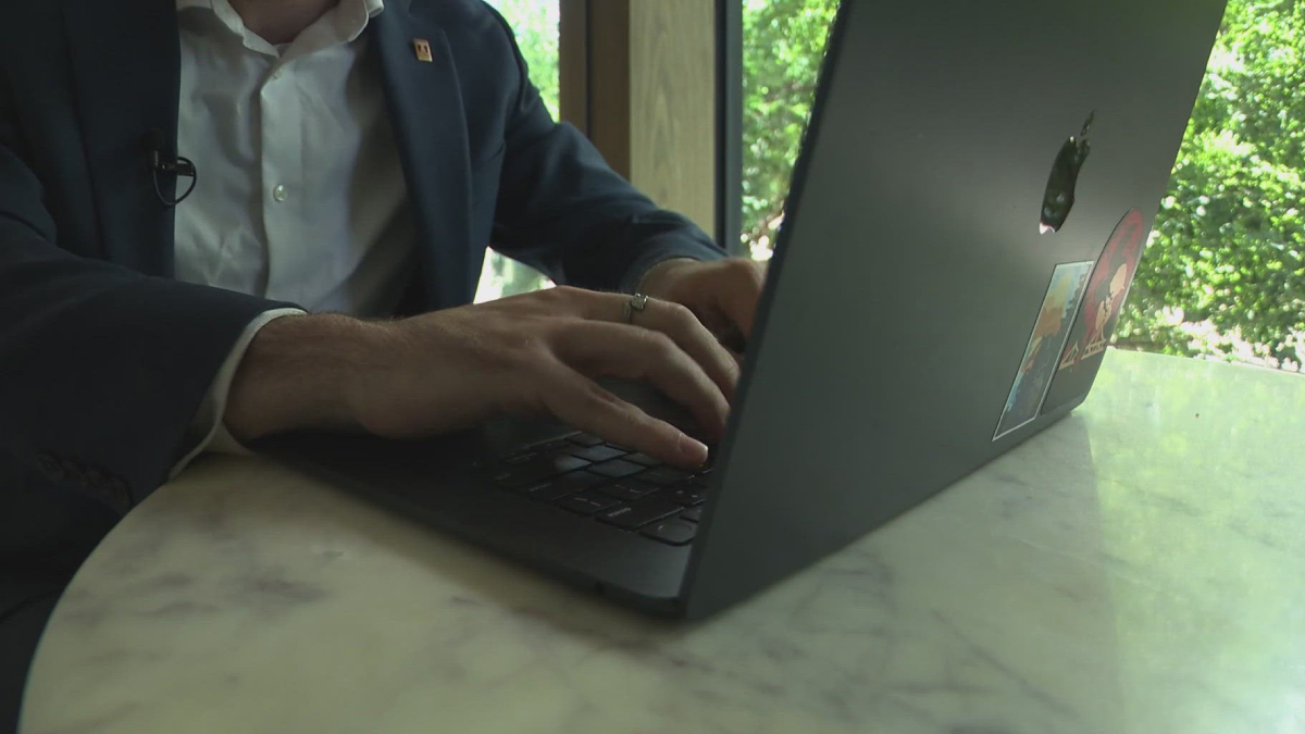 Person in a suit types on a MacBook with stickers at a marble table by a window with greenery outside