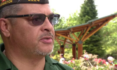 Close-up of a man wearing a Washington-cap and sunglasses, speaking outdoors in a flower-filled garden with a wooden pergola behind him.