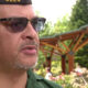 Close-up of a man wearing a Washington-cap and sunglasses, speaking outdoors in a flower-filled garden with a wooden pergola behind him.