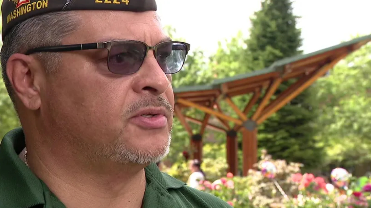Close-up of a man wearing a Washington-cap and sunglasses, speaking outdoors in a flower-filled garden with a wooden pergola behind him.