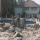 Rubble-filled backyard with a damaged brick structure and a curved swimming pool, a two-story house behind.