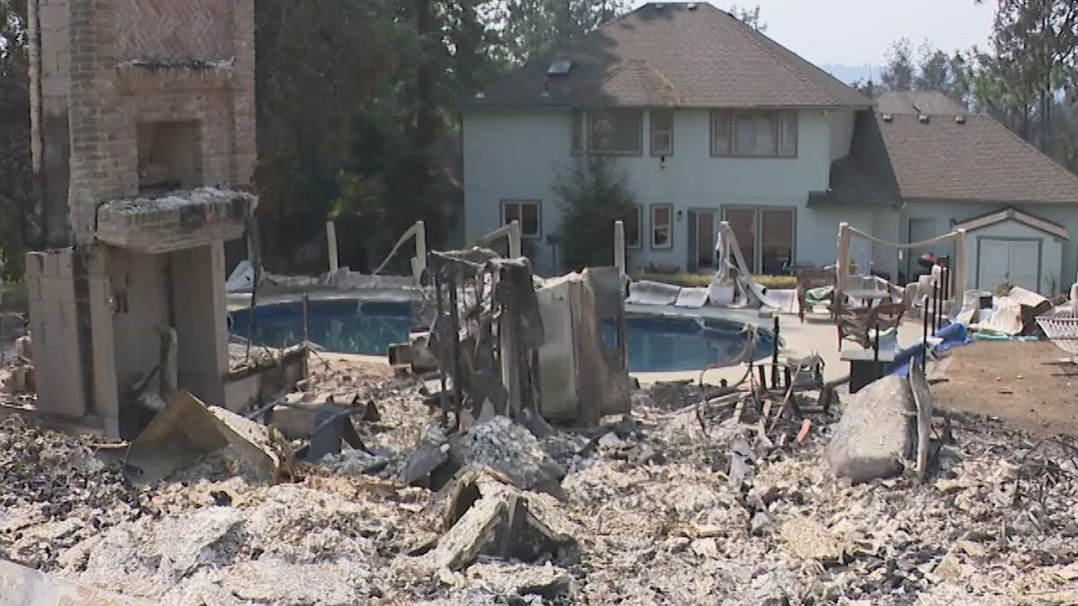 Rubble-filled backyard with a damaged brick structure and a curved swimming pool, a two-story house behind.