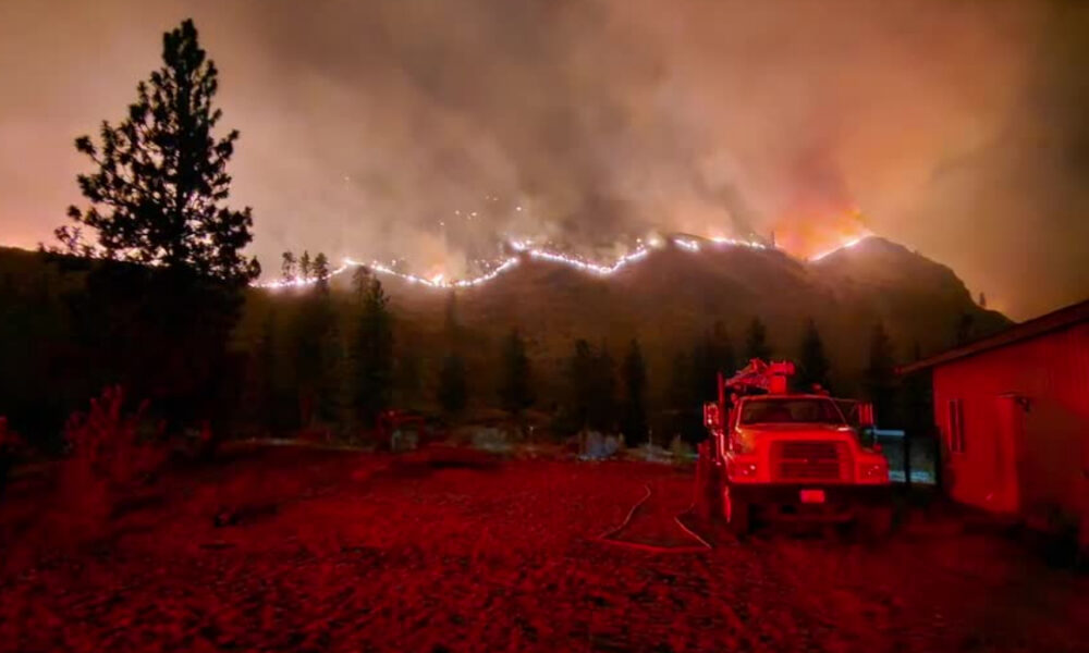 Night scene of a hillside wildfire with flames along the ridge, smoke in the sky, and a red fire truck parked beside a house in the foreground.