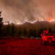 Night scene of a hillside wildfire with flames along the ridge, smoke in the sky, and a red fire truck parked beside a house in the foreground.