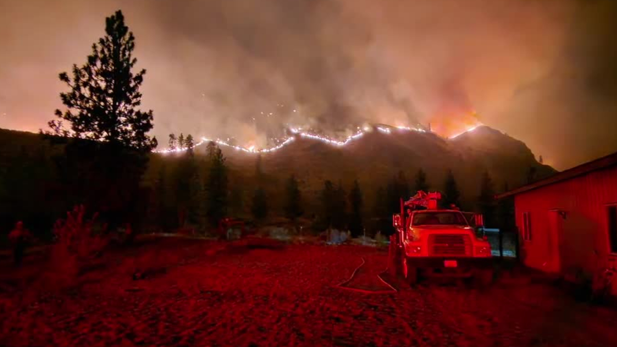Night scene of a hillside wildfire with flames along the ridge, smoke in the sky, and a red fire truck parked beside a house in the foreground.