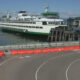 White passenger ferry docked at a pier with orange safety barriers along a curved road in the foreground.