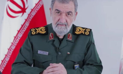 Senior male military officer in a green uniform with gold insignia sits at a desk, Iranian flag in the background.