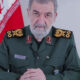 Senior male military officer in a green uniform with gold insignia sits at a desk, Iranian flag in the background.