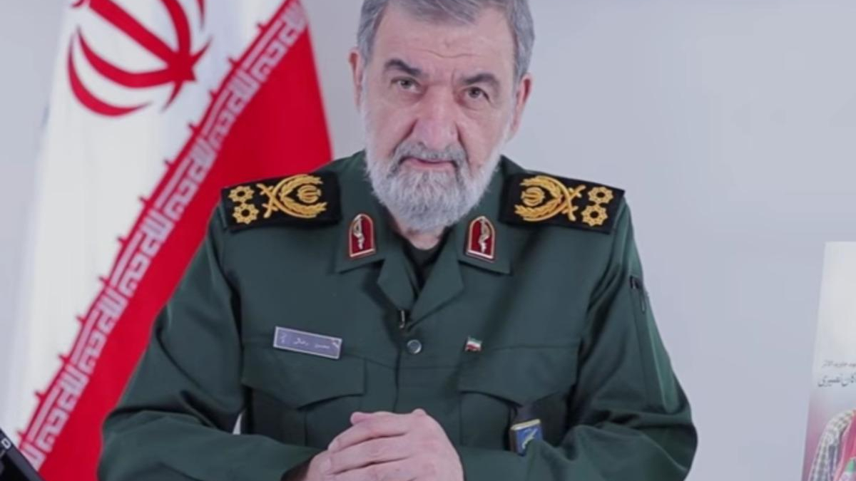 Senior male military officer in a green uniform with gold insignia sits at a desk, Iranian flag in the background.