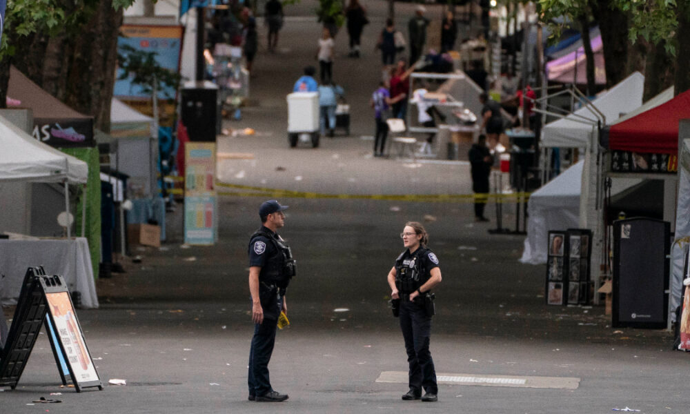 Two police officers standing and talking in the middle of a closed street market, with vendor tents and bystanders in the background.