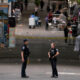 Two police officers standing and talking in the middle of a closed street market, with vendor tents and bystanders in the background.