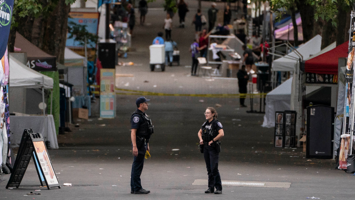 Two police officers standing and talking in the middle of a closed street market, with vendor tents and bystanders in the background.