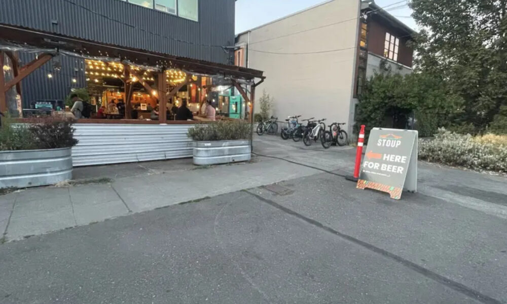 Outdoor restaurant patio with string lights and customers inside, bikes parked along the side, and a chalkboard sign outside offering beer