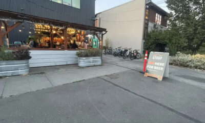 Outdoor restaurant patio with string lights and customers inside, bikes parked along the side, and a chalkboard sign outside offering beer