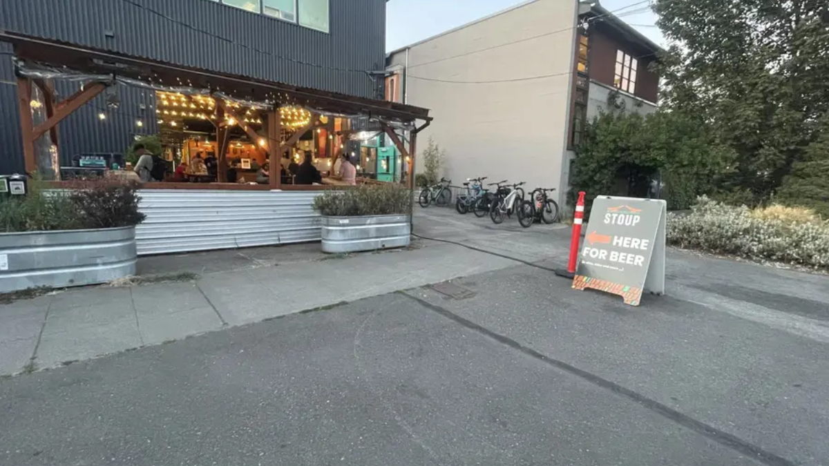 Outdoor restaurant patio with string lights and customers inside, bikes parked along the side, and a chalkboard sign outside offering beer