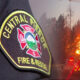 Close-up of a firefighter patch reading 'Central Paciﬁc Fire & Rescue' with a blazing house in the background, flames visible through trees, suggesting an active fire scene.