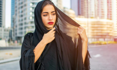 Woman in a black hijab outdoors in a city, adjusting her veil with a thoughtful expression.