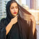 Woman in a black hijab outdoors in a city, adjusting her veil with a thoughtful expression.