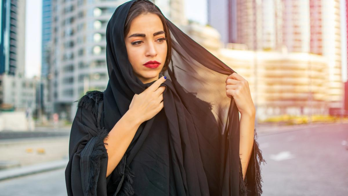 Woman in a black hijab outdoors in a city, adjusting her veil with a thoughtful expression.