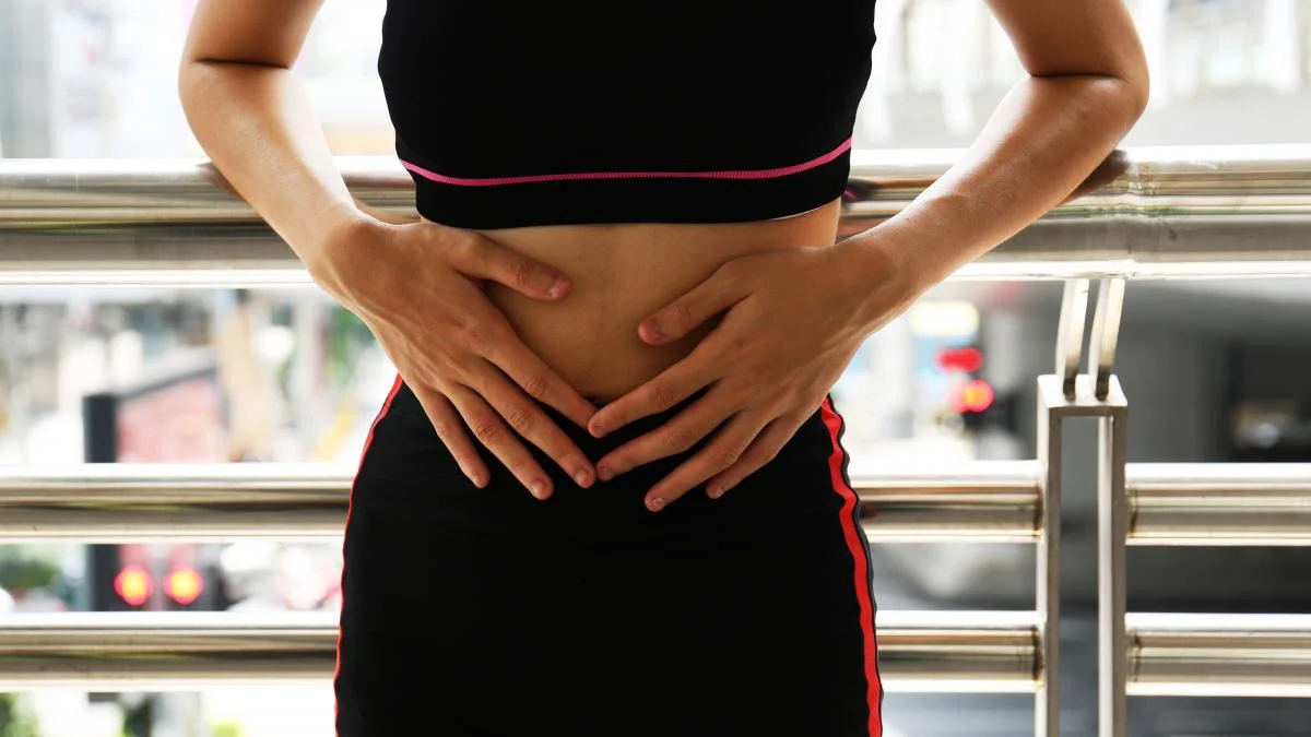 Person in a black athletic outfit holding their abdomen with both hands, gripping the midsection by a metal railing outdoors
