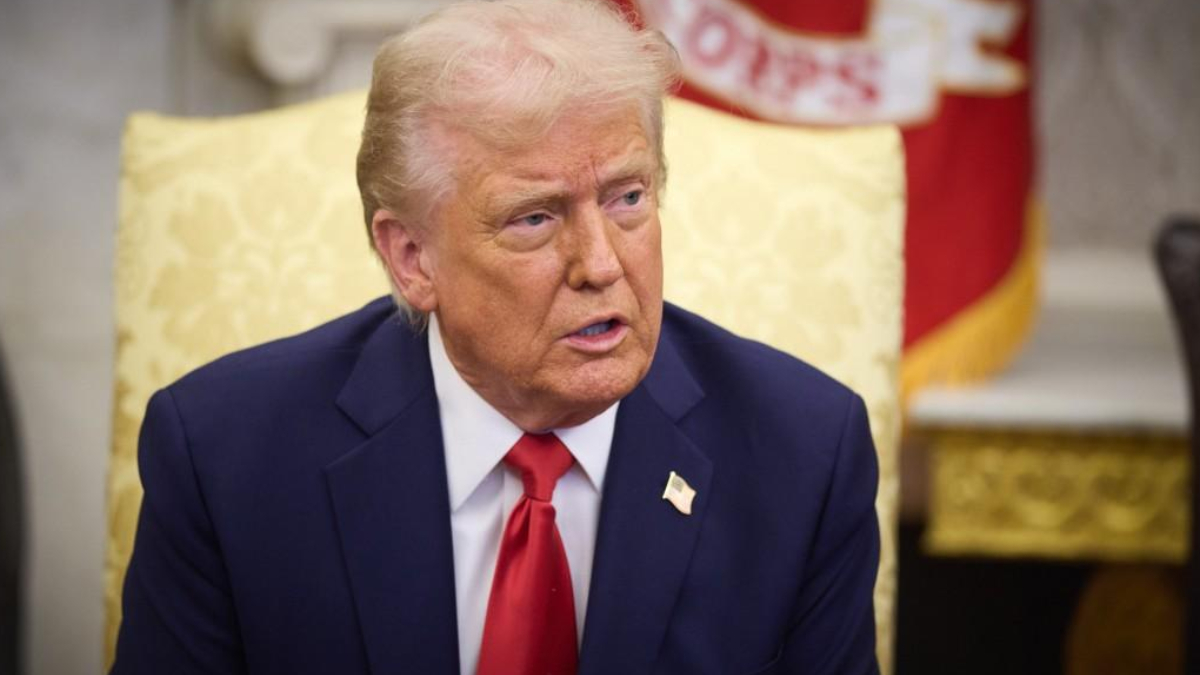 A man in a dark blue suit and red tie sits in a gold-patterned chair in a formal room, looking to the right with a serious expression.