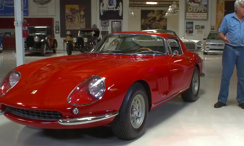 Red vintage Ferrari sports car on a showroom floor with a man in jeans gesturing beside it, inside a museum-like space with other classic cars in the background.