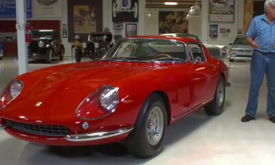 Red vintage Ferrari sports car on a showroom floor with a man in jeans gesturing beside it, inside a museum-like space with other classic cars in the background.