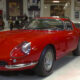 Red vintage Ferrari sports car on a showroom floor with a man in jeans gesturing beside it, inside a museum-like space with other classic cars in the background.