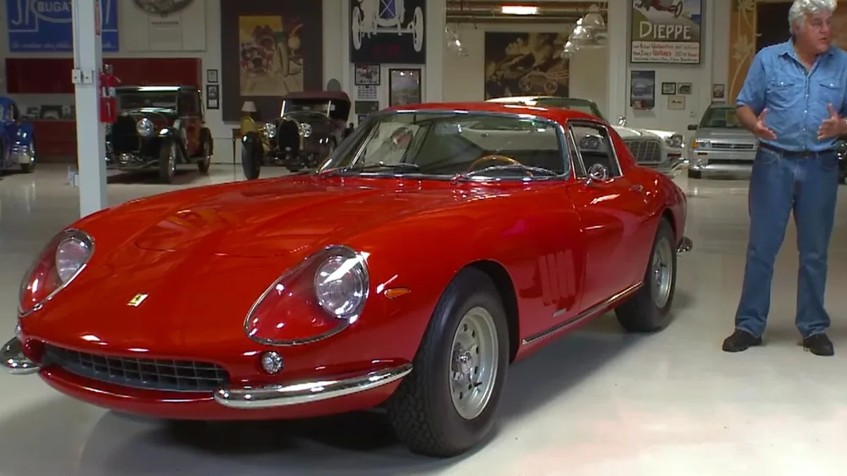Red vintage Ferrari sports car on a showroom floor with a man in jeans gesturing beside it, inside a museum-like space with other classic cars in the background.