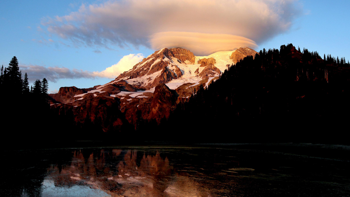 Snow-capped mountain at sunset with a lenticular cloud above, flanked by dark forest and a reflecting lake in the foreground.