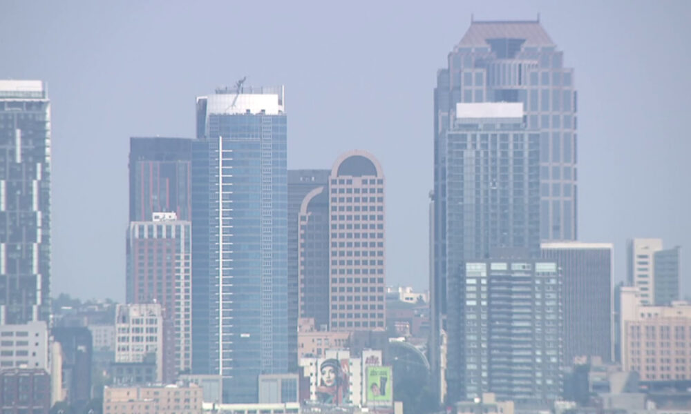 Blue-tinted city skyline with several tall glass office towers against a hazy sky.