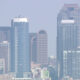 Blue-tinted city skyline with several tall glass office towers against a hazy sky.