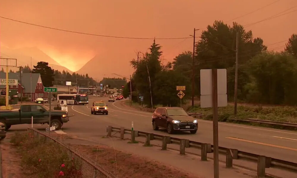 Roadway in a small town at sunset with thick orange smoke in the sky and distant mountains; several vehicles on the street.