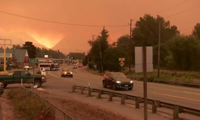 Roadway in a small town at sunset with thick orange smoke in the sky and distant mountains; several vehicles on the street.