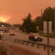 Roadway in a small town at sunset with thick orange smoke in the sky and distant mountains; several vehicles on the street.