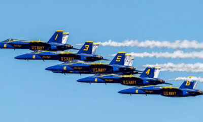 Six Blue Angels jets fly in a tight diagonal formation with white smoke trails against a clear blue sky, tails numbered 6, 4, 5, 3, and 9 visualized on blue fuselages.