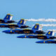 Six Blue Angels jets fly in a tight diagonal formation with white smoke trails against a clear blue sky, tails numbered 6, 4, 5, 3, and 9 visualized on blue fuselages.
