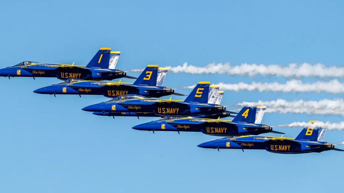 Six Blue Angels jets fly in a tight diagonal formation with white smoke trails against a clear blue sky, tails numbered 6, 4, 5, 3, and 9 visualized on blue fuselages.