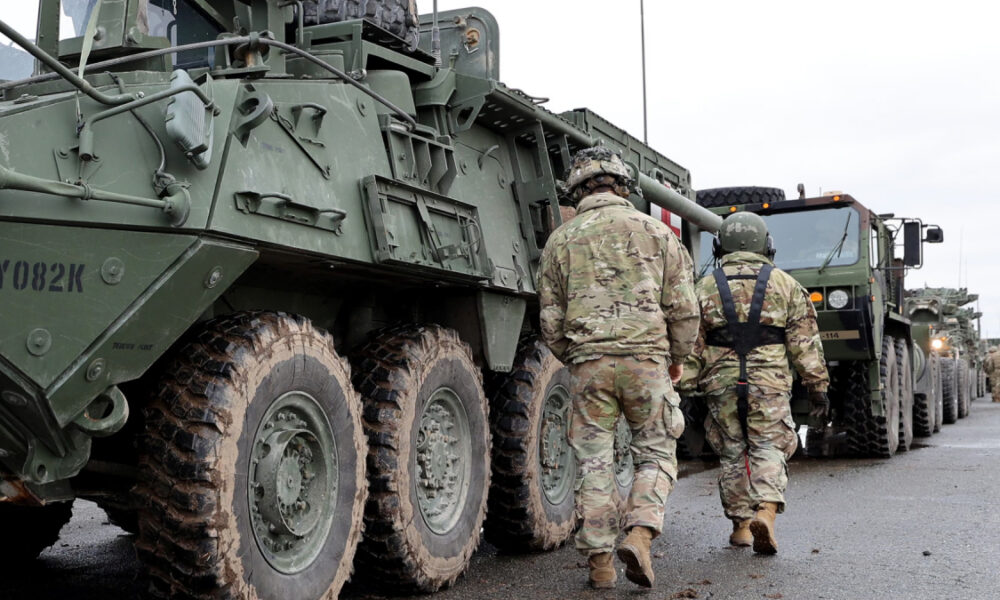 Soldiers in camouflage walk beside a row of large military armored vehicles on a paved road, preparing for a convoy move