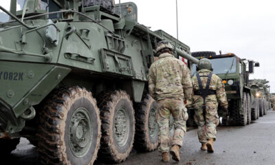 Soldiers in camouflage walk beside a row of large military armored vehicles on a paved road, preparing for a convoy move