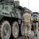 Soldiers in camouflage walk beside a row of large military armored vehicles on a paved road, preparing for a convoy move