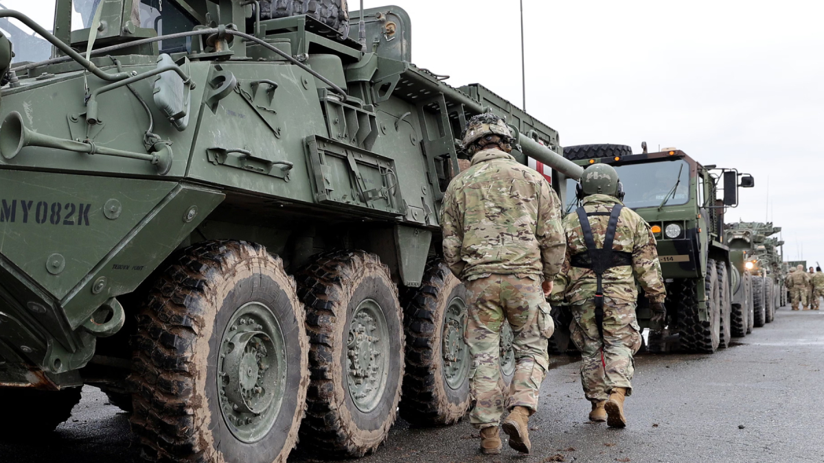 Soldiers in camouflage walk beside a row of large military armored vehicles on a paved road, preparing for a convoy move