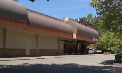 Exterior of a shopping center with a large curved brown awning and parked cars along the curb under a blue sky.