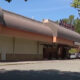 Exterior of a shopping center with a large curved brown awning and parked cars along the curb under a blue sky.