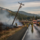 Firefighters spraying a grass fire along a wet highway, a leaning power pole toppled beside the flames, smoke rising with forested hills beyond.