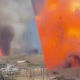 Industrial site on fire with large orange flames and thick black smoke rising from buildings.
