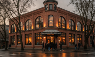 Corner brick building with large arched windows glowing warmly as people enter from a wet street at dusk.