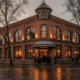 Corner brick building with large arched windows glowing warmly as people enter from a wet street at dusk.